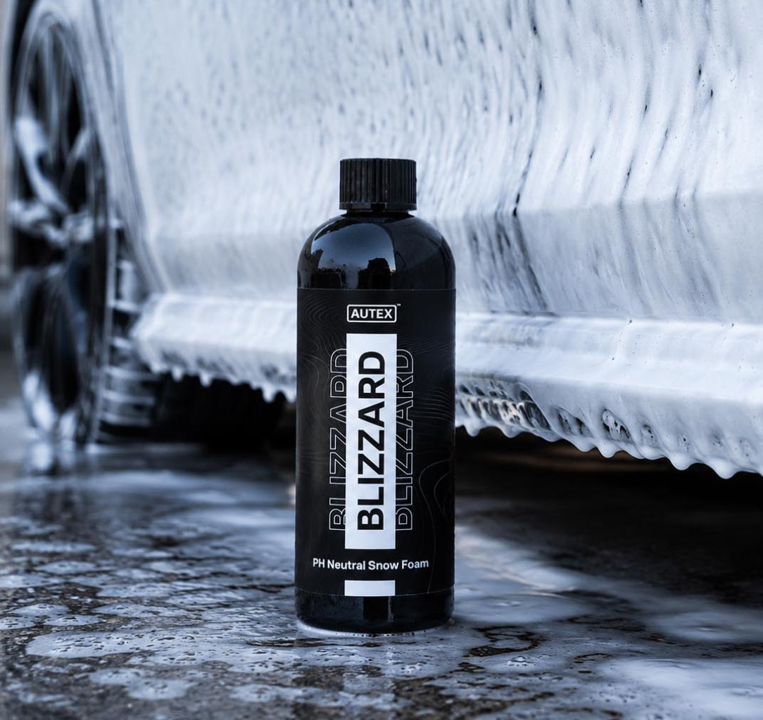 A bottle of Autex Blizzard Blazing pH Natural Snow Foam stands on wet pavement in front of a car covered in white snow foam.